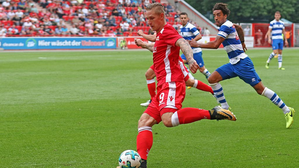 A soccer player in a red jersey kicks the ball while a player in a striped jersey chases him.
