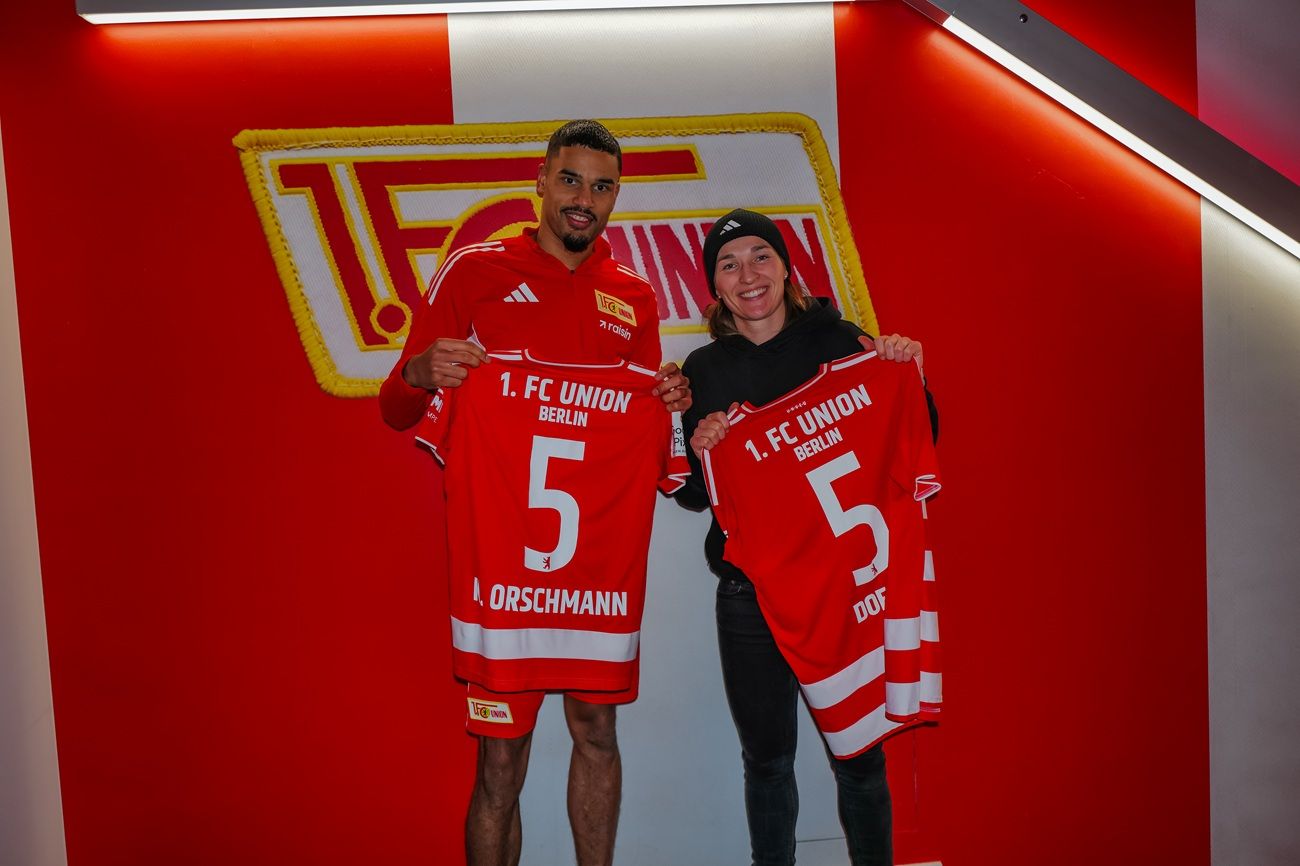 Zwei Personen halten jeweils ein rotes Trikot mit der Aufschrift "1. FC UNION" und der Nummer 5 in einer stadionähnlichen Umgebung.