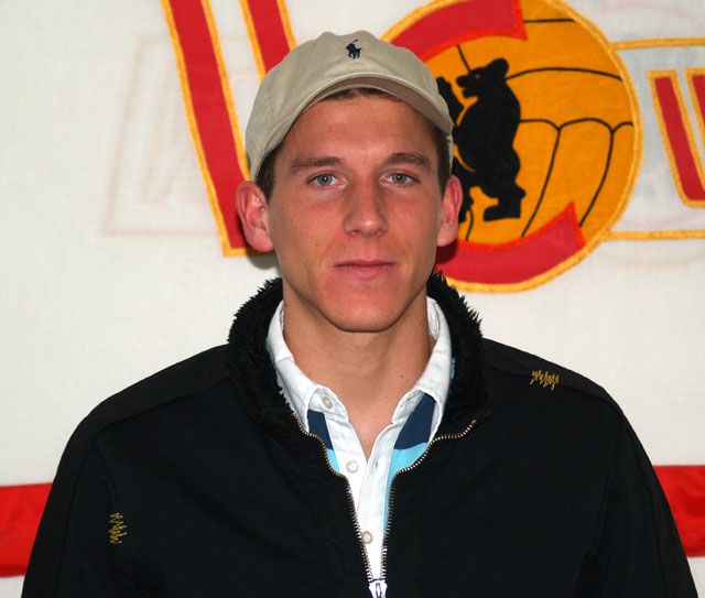 Ein junger Mann mit einem beigen Hut und schwarzer Jacke steht vor einer bunten Wand mit Sportmotiven.