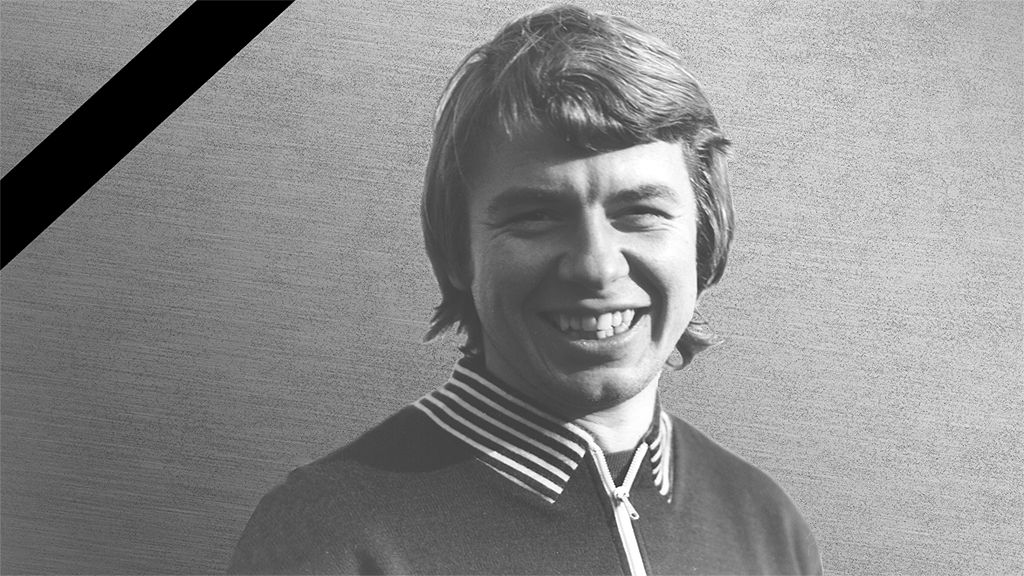 Schwarz-weiß Porträt eines lächelnden Mannes mit mittellangen, lockigen Haaren und einem Sportpullover mit Kragen.