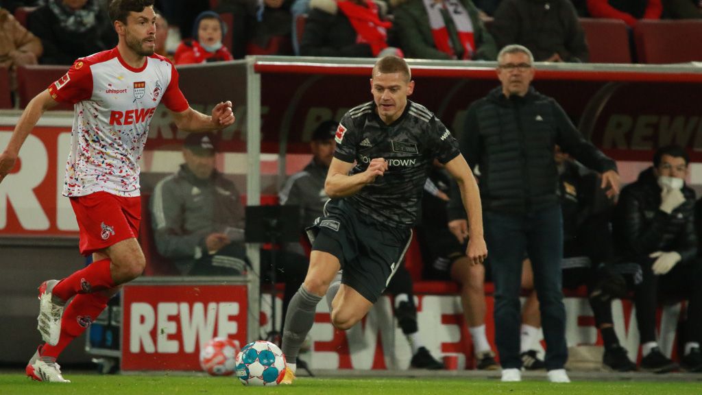 A football player in a gray jersey dribbles the ball while an opponent chases him. In the background, spectators can be seen.