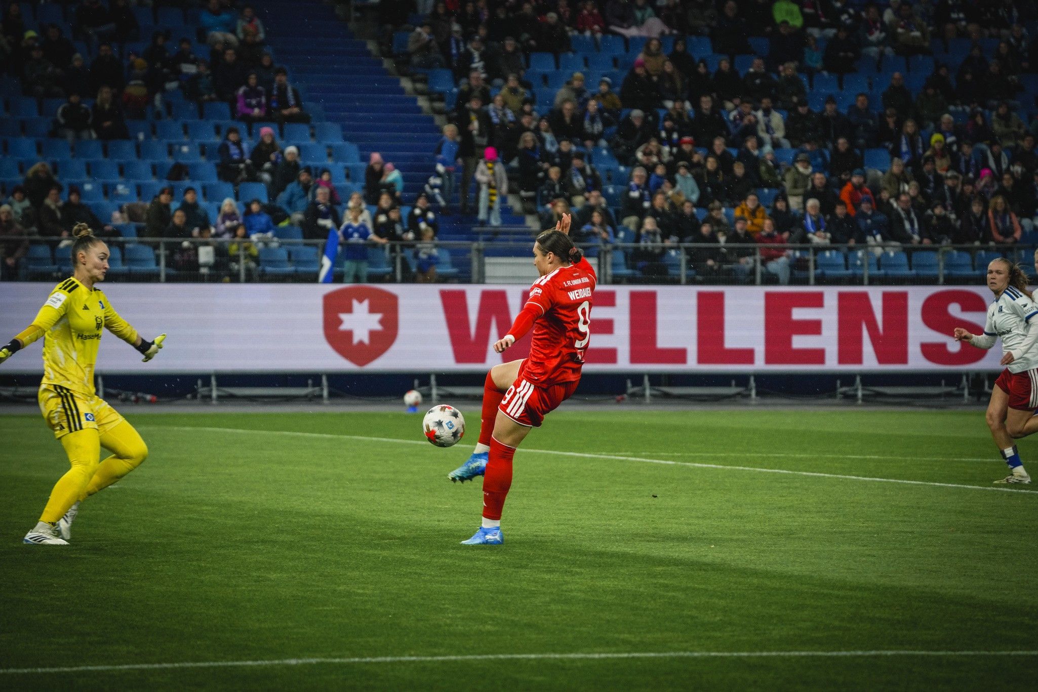 Eine Spielerin in rotem Trikot schießt den Ball ins Tor, während die Torhüterin in gelbem Trikot reagiert. Im Hintergrund Publikum und Werbebanner.