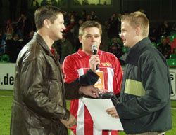 Drei Männer stehen auf einem Sportfeld, einer hält ein Mikrofon, die anderen zwei sprechen miteinander, einer trägt ein Fußballtrikot.