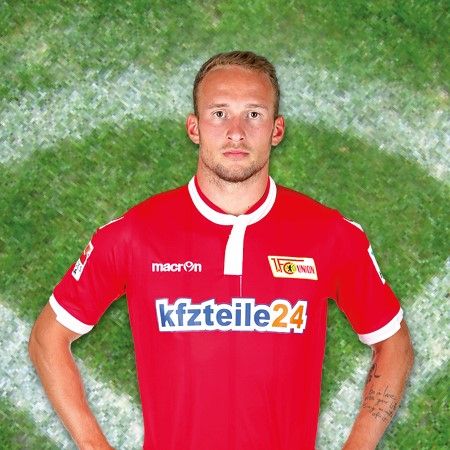 Person in rotem Trikot mit Sponsorenlogo vor grünem Fußballfeld-Hintergrund.