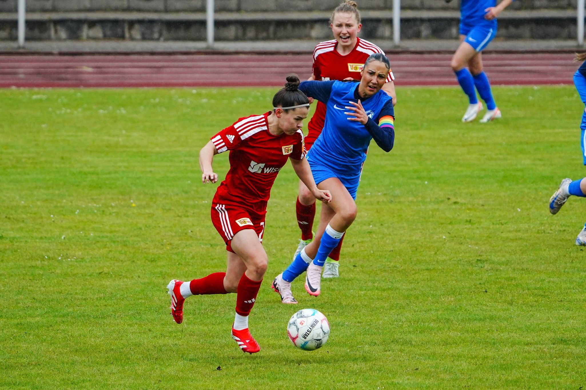 Eine Spielerin in rotem Trikot dribbelt den Ball, während eine Gegnerin in blauem Trikot versucht, den Ball zu erobern.