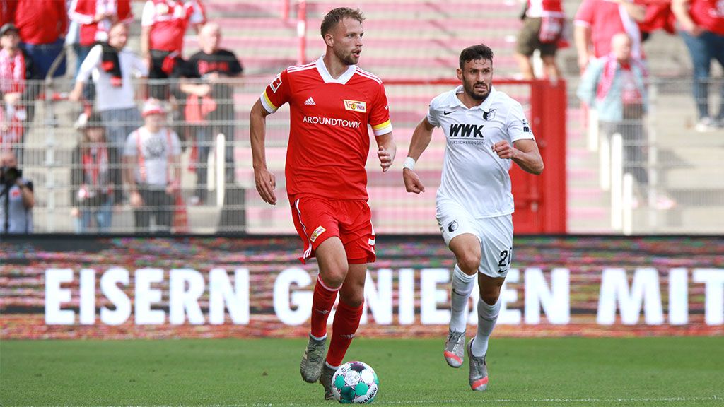 Ein Fußballspieler in rotem Trikot dribbelt den Ball, während ein Gegner in weißem Trikot ihn verfolgt. Stadion im Hintergrund.