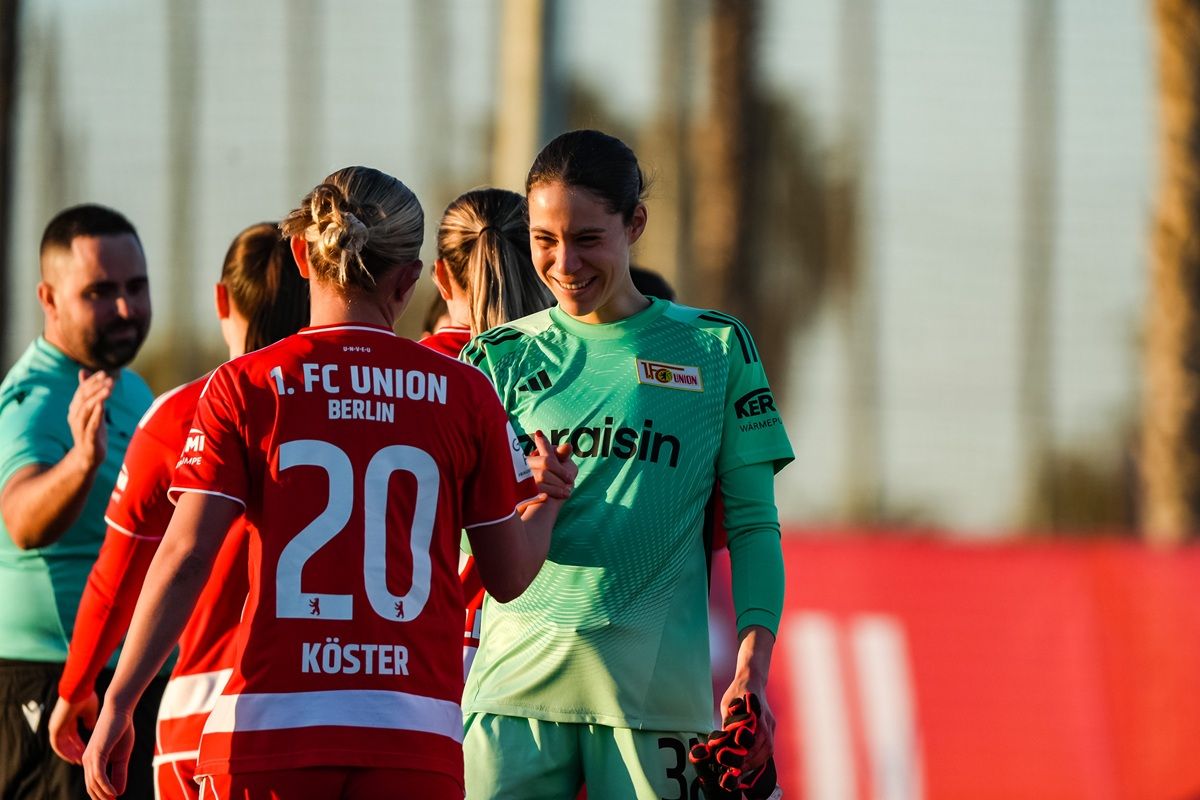 Zwei Fußballspielerinnen, eine in rotem Trikot und eine in grüner Torwartausrüstung, lächeln sich während des Aufeinandertreffens zu.