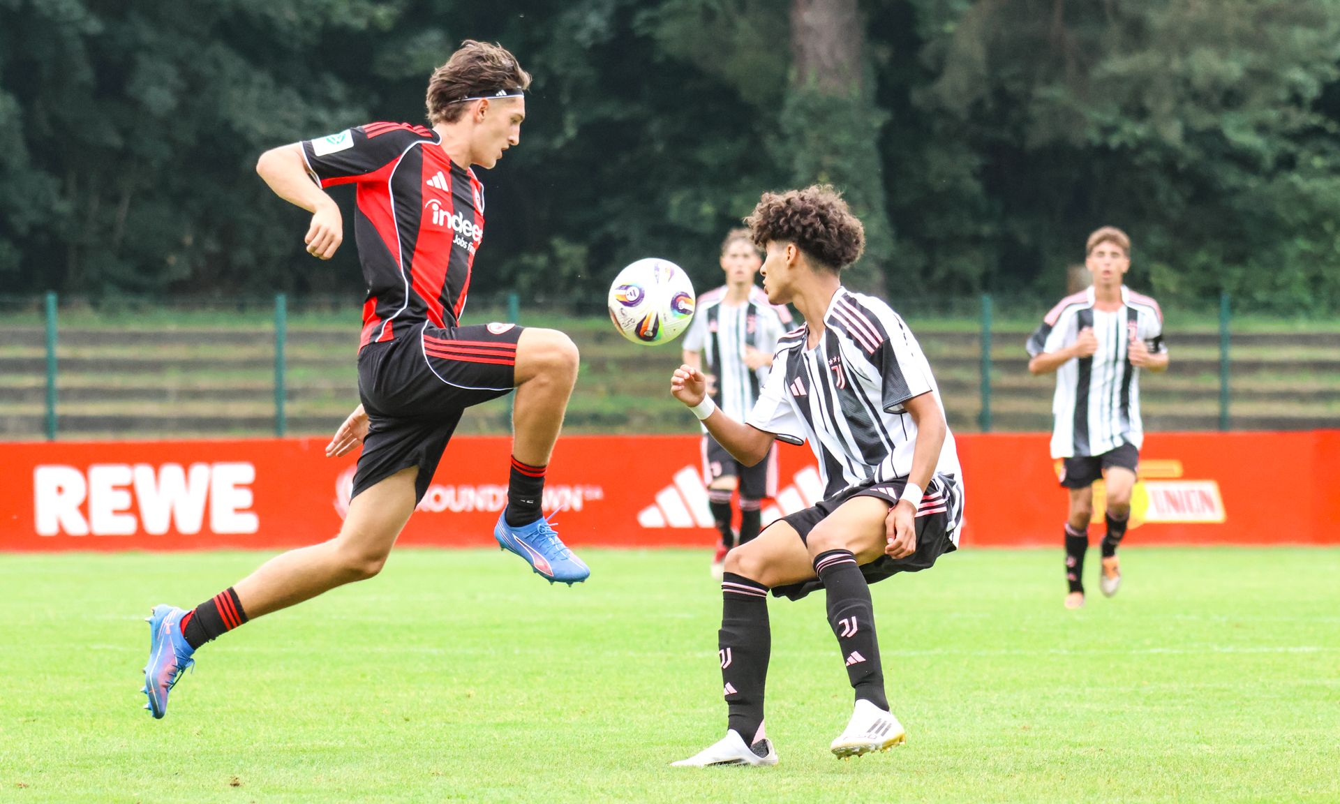 Ein Fußballspieler springt über einen gegnerischen Spieler auf einem Rasenplatz, während andere Spieler im Hintergrund zuschauen.