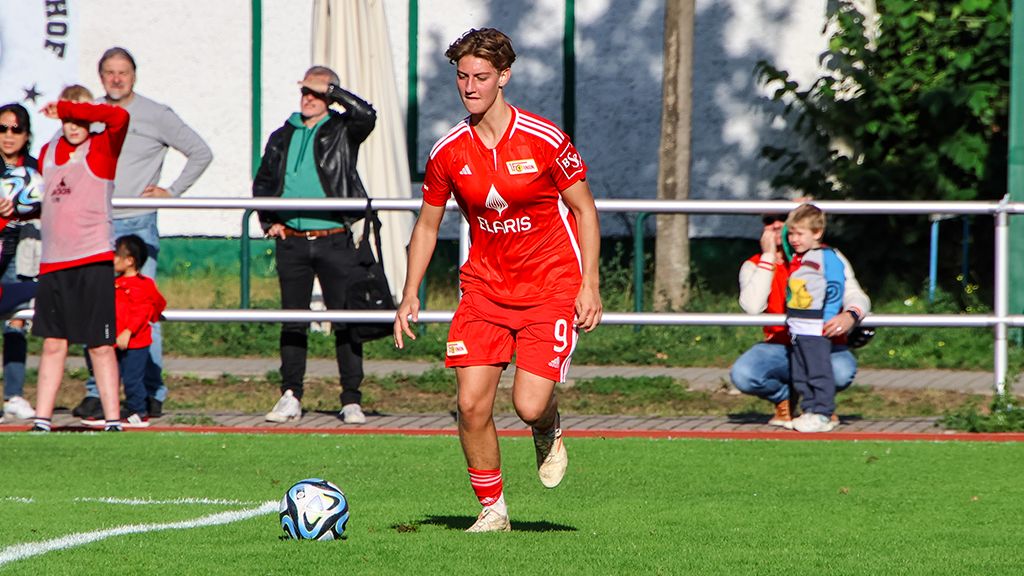 A female soccer player in a red jersey is running with the ball on a grass field while spectators watch in the background.