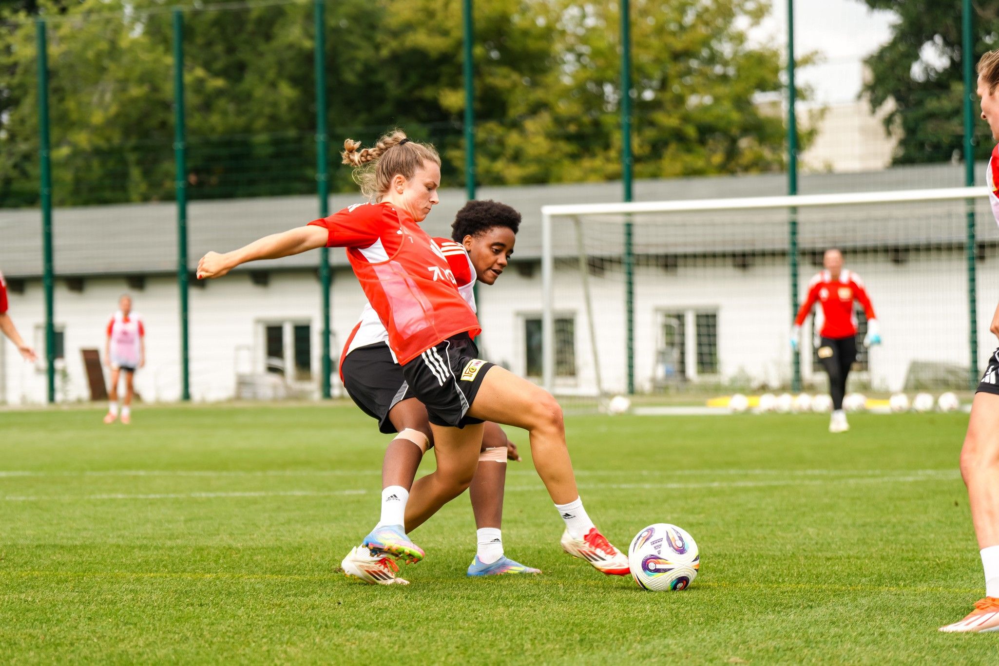 Zwei Fußballerinnen in roten Trikots kämpfen um den Ball auf einem grünen Trainingsplatz.