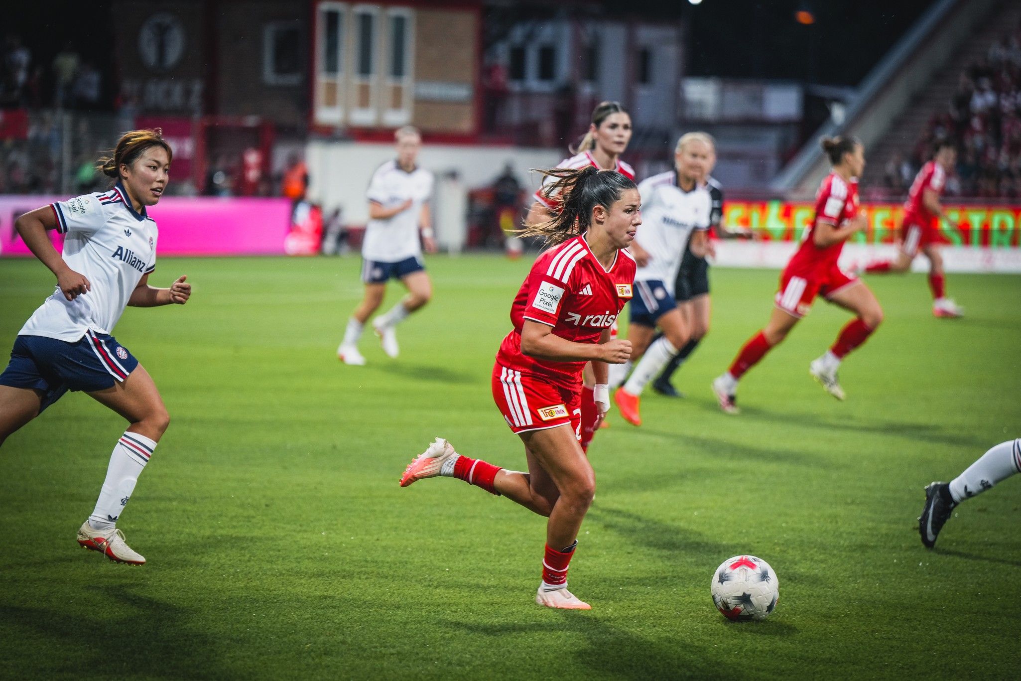 Spielszene im Fußball mit zwei Mannschaften auf dem Platz, eine Spielerin in Rot dribbelt den Ball während andere Spielerinnen verfolgen.