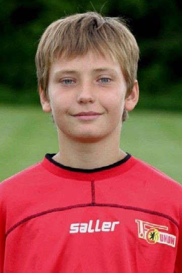 Ein junger Fußballspieler mit kurzen, blonden Haaren trägt ein rotes Trikot und lächelt in die Kamera.
