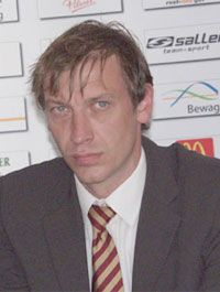 Ein Mann mit ungekämmten Haaren trägt einen Anzug und sitzt an einem Tisch mit Logos im Hintergrund.