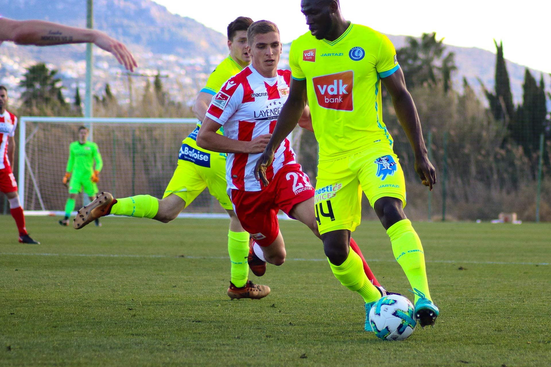 Zwei Fußballspieler spielen auf einem grünen Platz. Einer trägt helle Farben. Der dritte Spieler trägt Rot. Er versucht, den Ball zu kriegen.