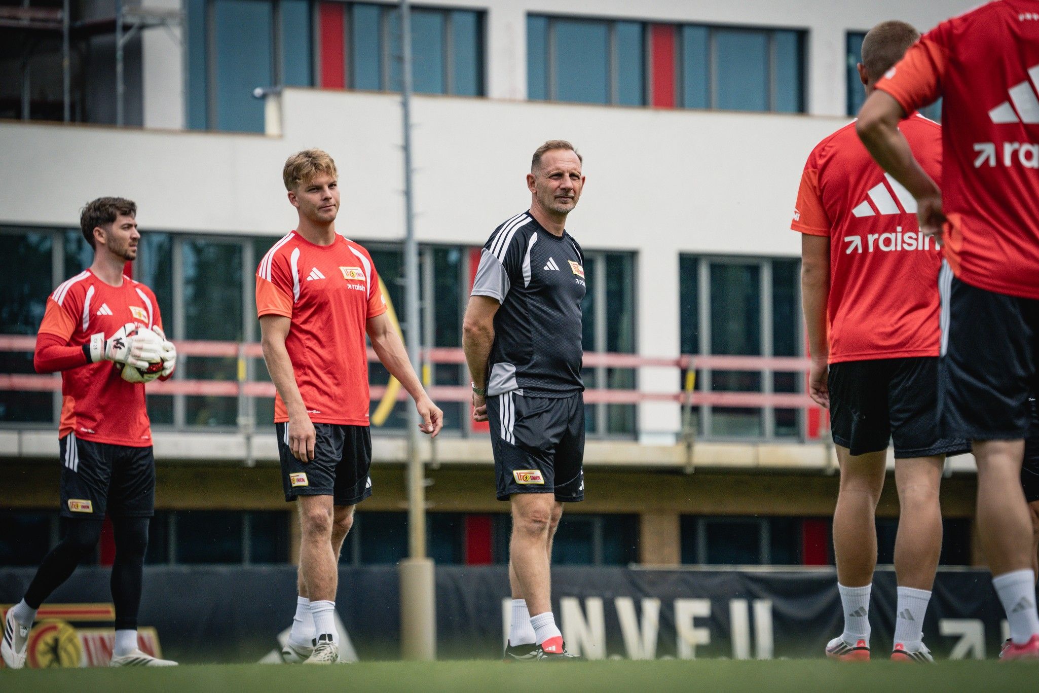 Eine Gruppe von Fußballspielern in roten Trikots trainiert auf dem Platz, während ein Trainer ihnen Anweisungen gibt.