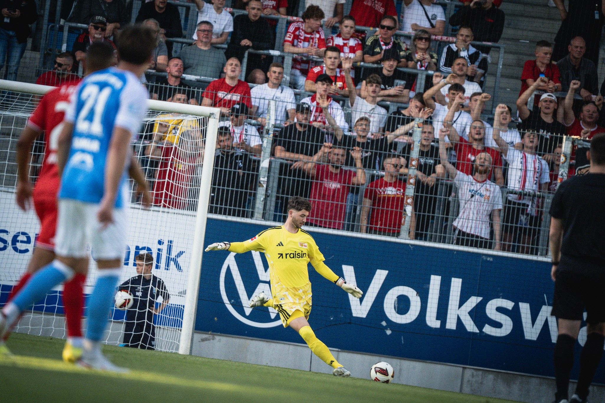 Fußballspiel mit einem Torwart in gelber Ausrüstung, der den Ball tritt, während Fans im Hintergrund jubeln.