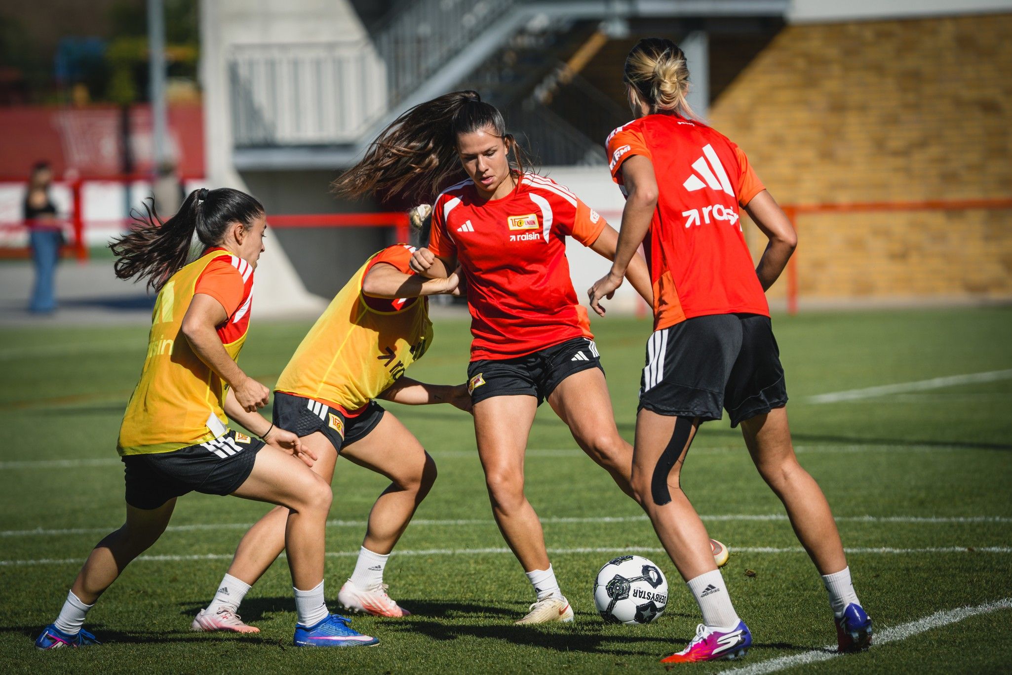 Eine Gruppe von vier weiblichen Fußballspielern trainiert auf einem grünen Platz. Drei Spielerinnen drängen sich um den Ball.