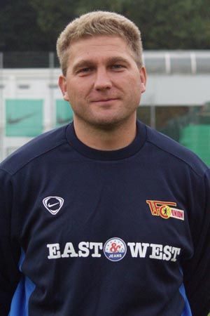 Ein Mann in einem Sportanzug mit einem Logo auf der Brust steht vor einer Sportanlage.