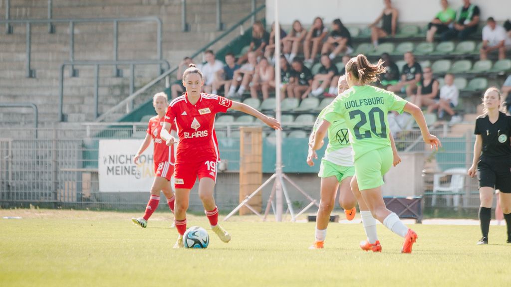 Two female soccer players competing on a sports field; one in red, the other in green. Spectators in the background.