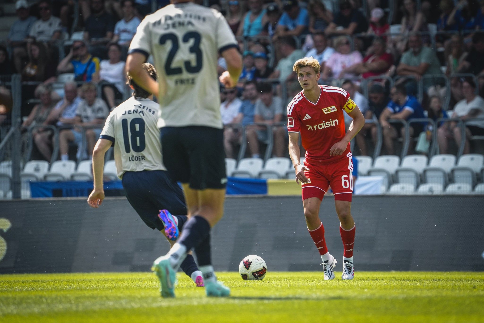 Spielszene im Fußball mit Spielern in roten und blauen Trikots, Zuschauer im Hintergrund. Der Ball ist im Spiel.