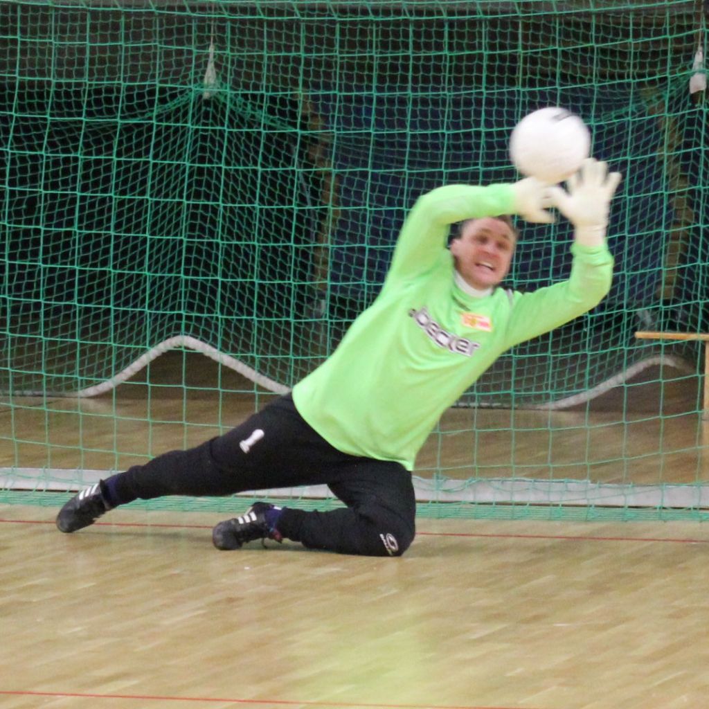 Torwart in grünem Trikot springt, um einen Ball zu fangen, vor einem Tor mit Netz im Hintergrund.