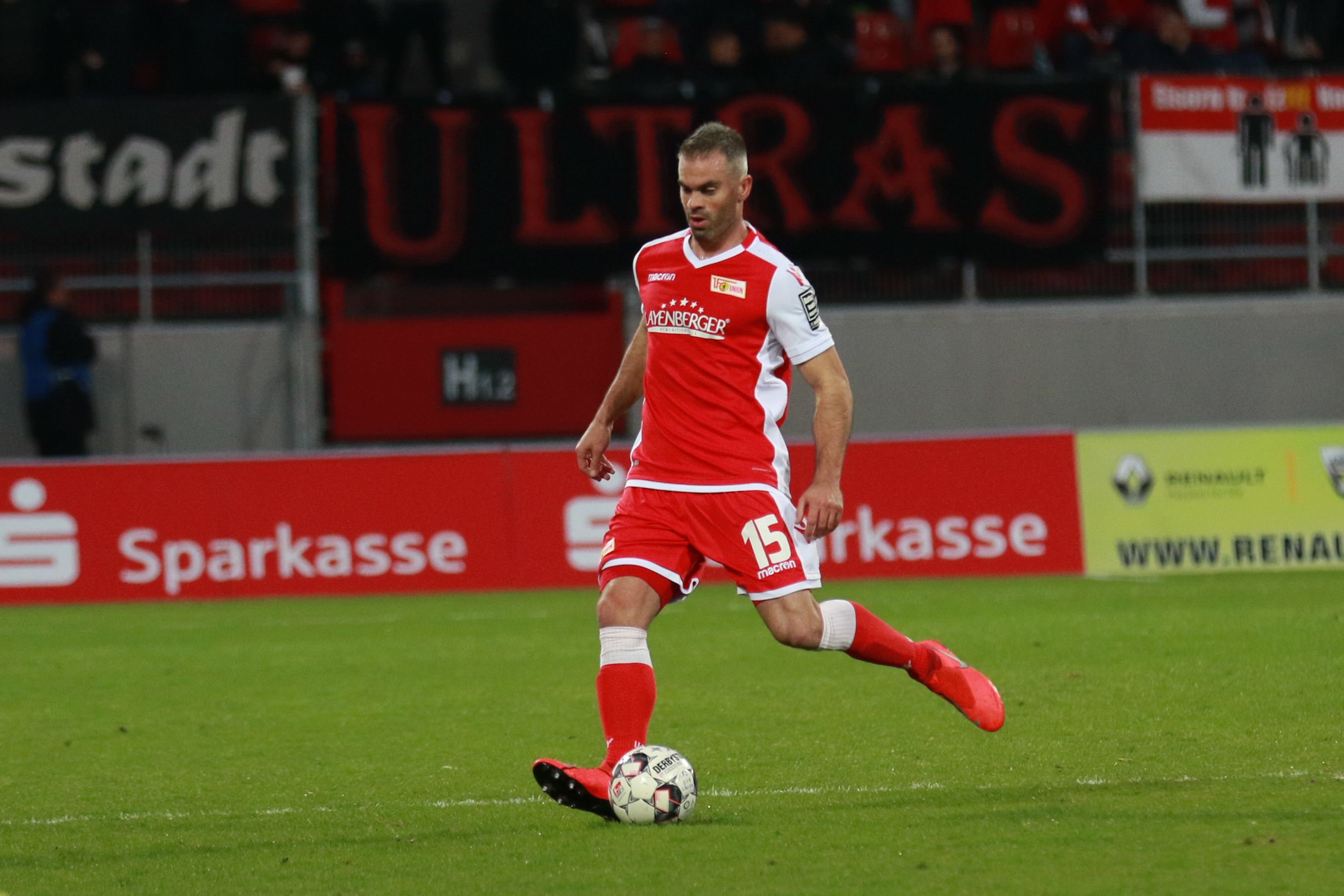 Un jugador de fútbol con uniforme rojo patea el balón en un campo. Al fondo hay aficionados y pancartas.