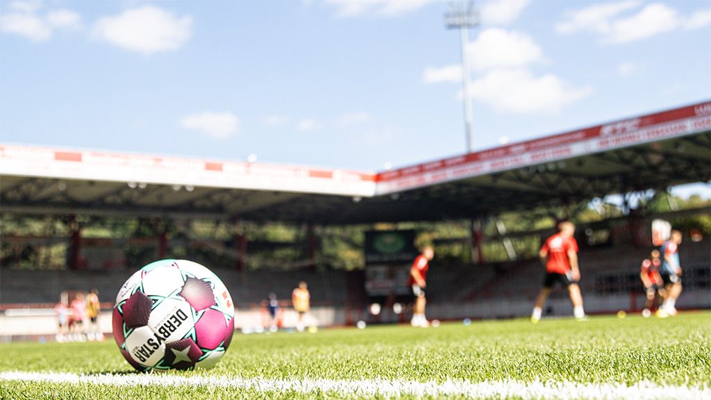 Ein Fußball liegt auf dem Rasen eines Stadions, im Hintergrund trainieren Spieler bei sonnigem Wetter.