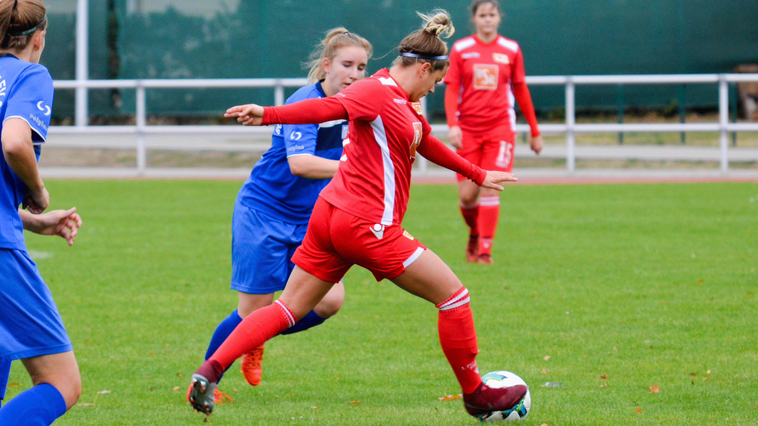 Eine Fußballspielerin trägt ein rotes Trikot. Sie dribbelt den Ball. Eine Spielerin in blauem Trikot kommt näher.