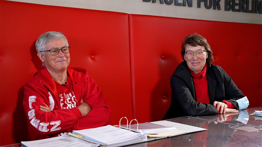 Zwei Personen sitzen an einem Tisch mit roten Wänden, lächeln und tragen Brillen. Unterlagen liegen auf dem Tisch.