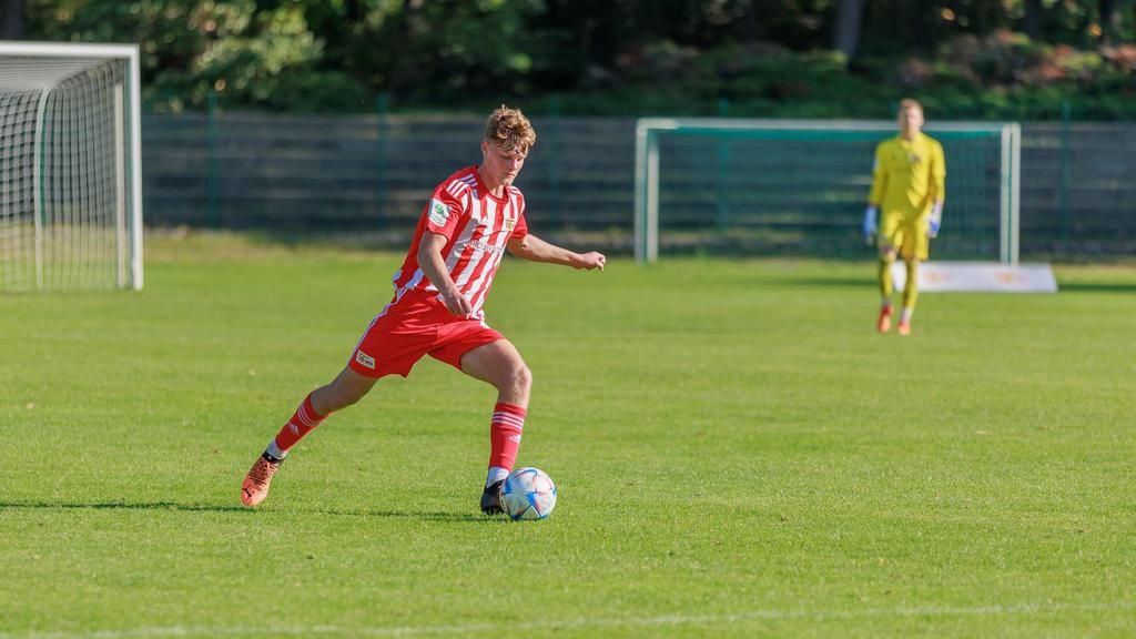 Ein Fußballspieler in rot-weißer Uniform schießt den Ball auf einem grünen Platz, während ein Torwart im Hintergrund steht.