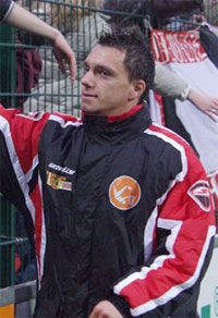 Ein Mann in einem schwarzen und roten Sportjacke steht in einer Sportarena und schaut interessiert in die Menge.