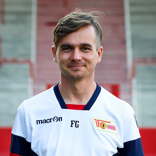 Ein lächelnder Mann in einem weißen Fußballtrikot mit roten und blauen Details steht vor einer Sportarena.