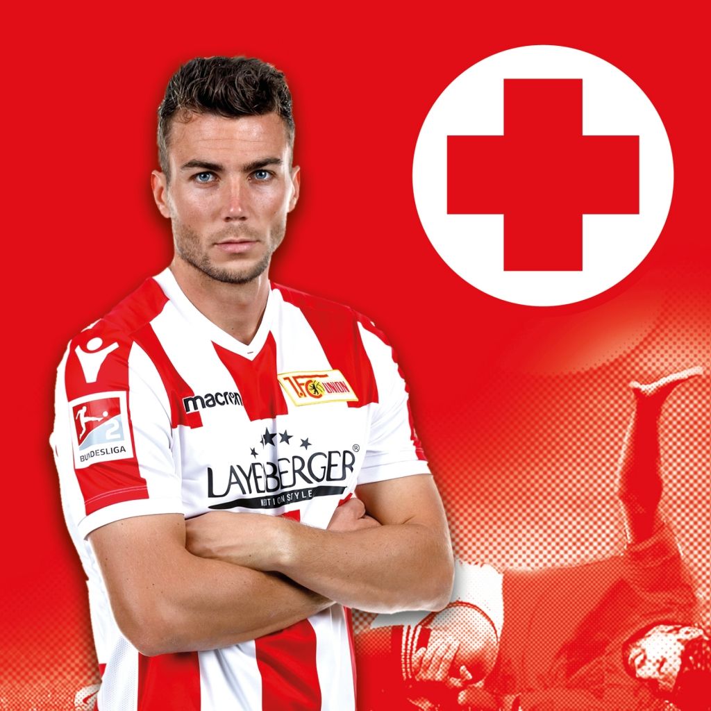 Ein Fußballspieler in Rot-Weißer Vereinskleidung mit einem roten Kreuz-Symbol im Hintergrund.