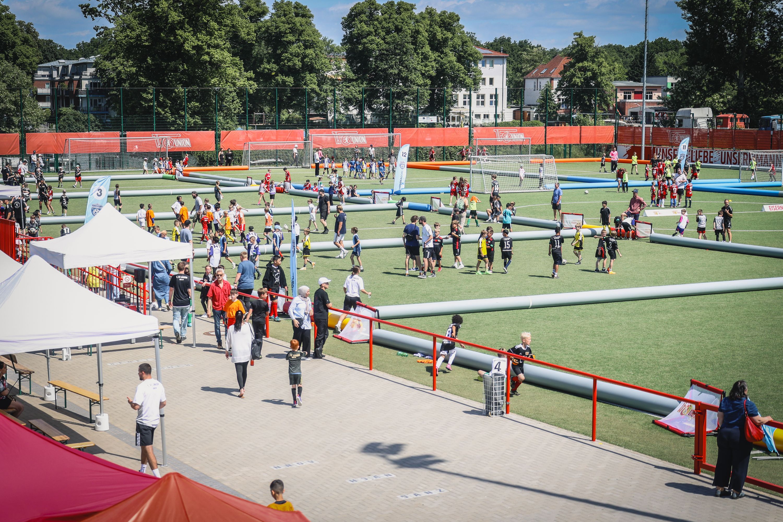 Auf einem Sportplatz spielen viele Menschen in bunten Trikots Fußball, umgeben von Zelten und Zuschauerbereichen.