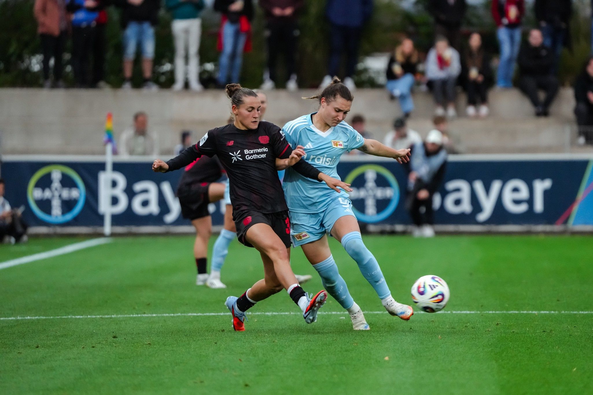 Zwei Frauenfußballspielerinnen kämpfen um den Ball auf einem Feld, umgeben von Zuschauern.