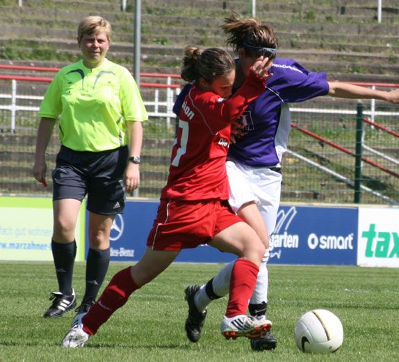 Aktive Fußballszene mit zwei Spielerinnen im Zweikampf, einer Schiedsrichterin im Hintergrund auf einem Spielfeld.
