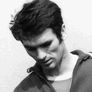 Ein Mann mit dunklen Haaren und schmalem Gesicht, der nach unten schaut und ein einfaches Oberteil trägt.