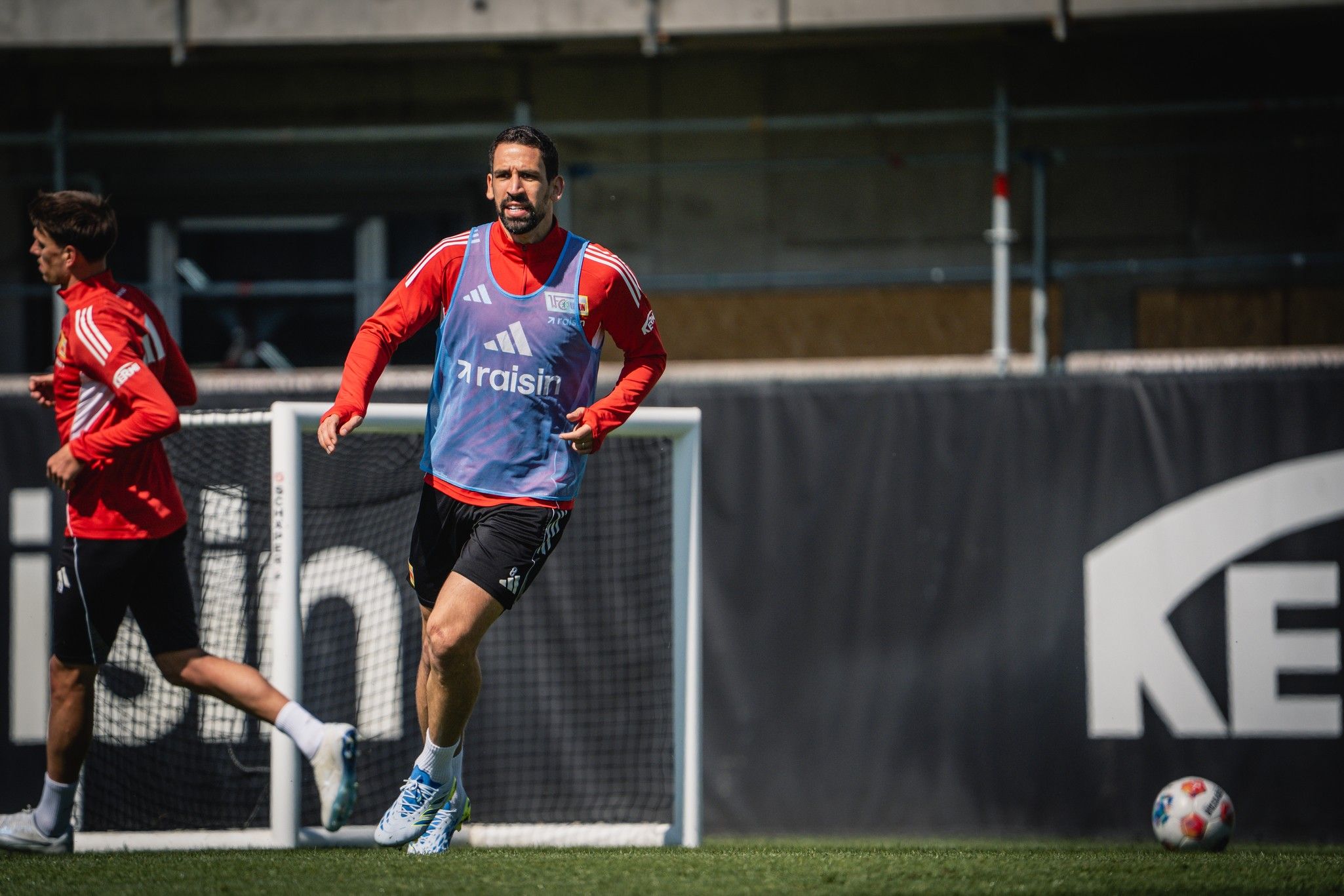 Ein Fußballspieler in Trainingskleidung läuft auf dem Platz, während ein weiterer Spieler im Hintergrund übt.
