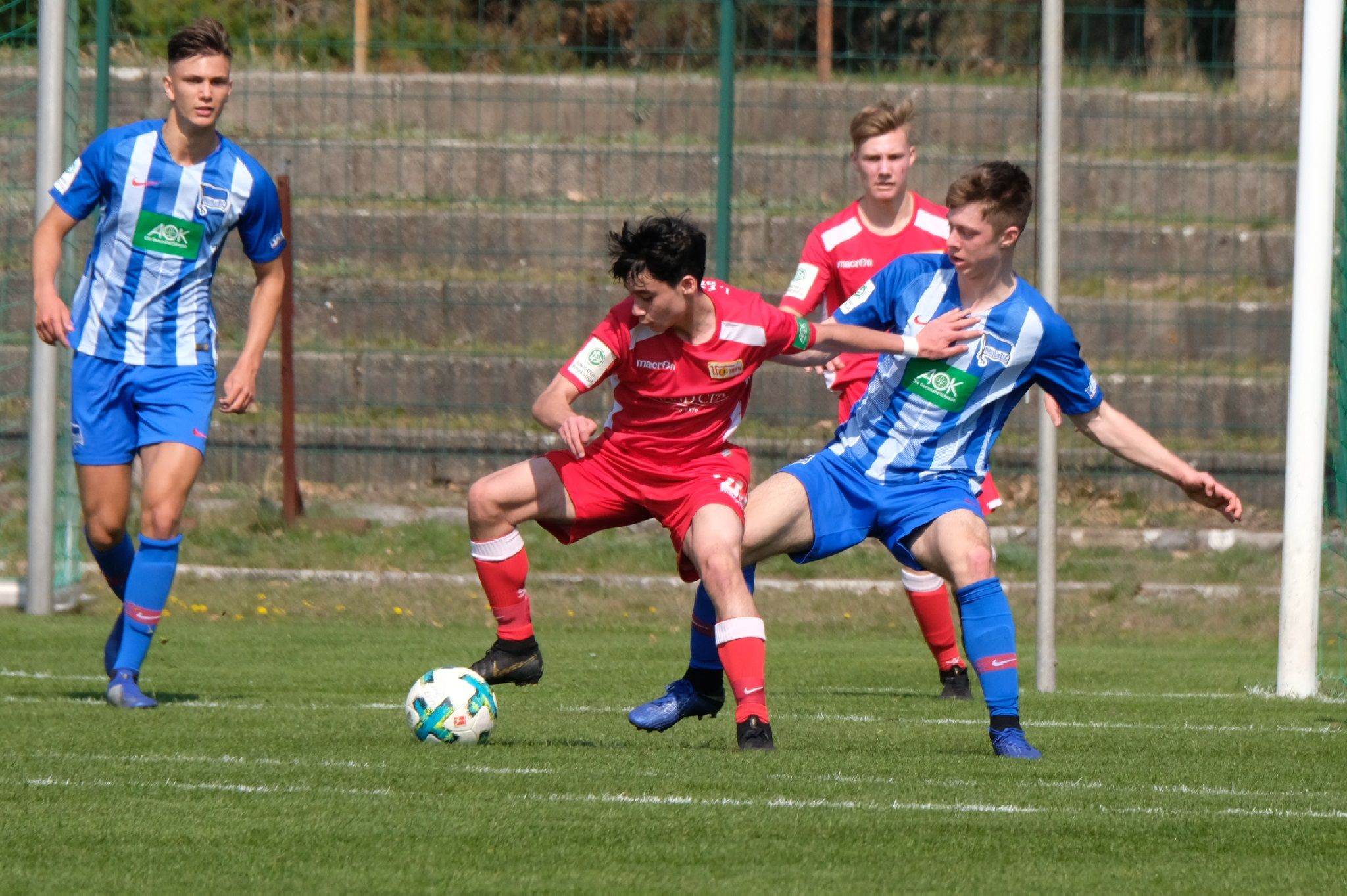 Zwei Fußballspieler kämpfen um den Ball, während ein weiterer Spieler in der Nähe zuschaut. Einheitliche Trikots in Rot und Blau.