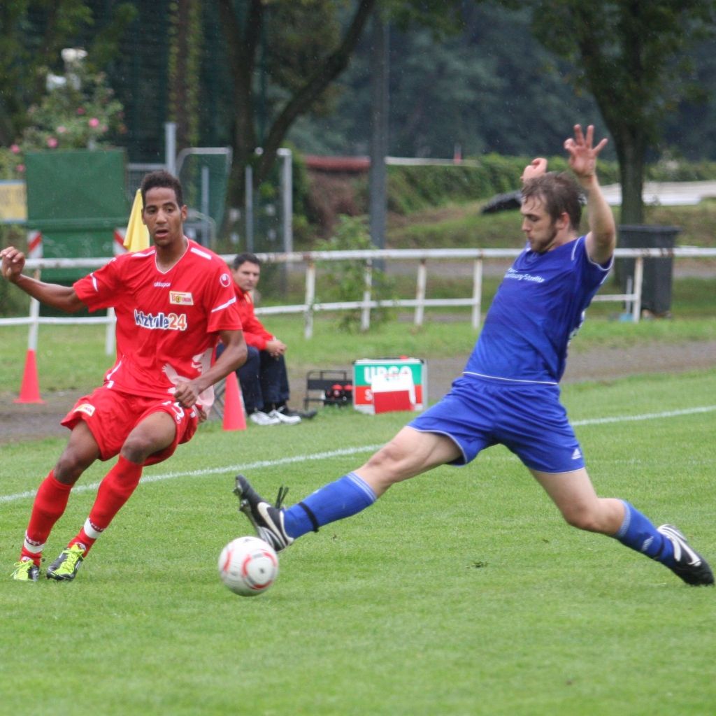 Zwei Fußballspieler im Wettkampf auf dem Spielfeld, einer in rot, der andere in blau, während ein Ball im Spiel ist.