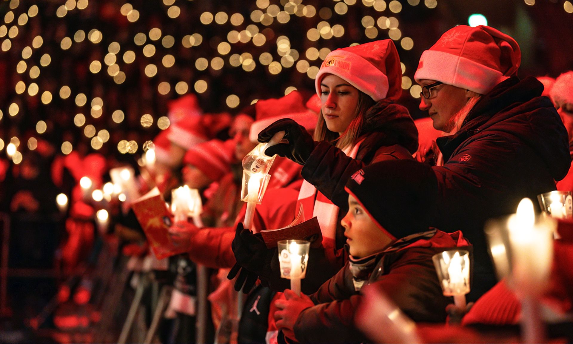 Menschen in Weihnachtsmützen halten Kerzen und Geschenke, umgeben von festlicher Beleuchtung.