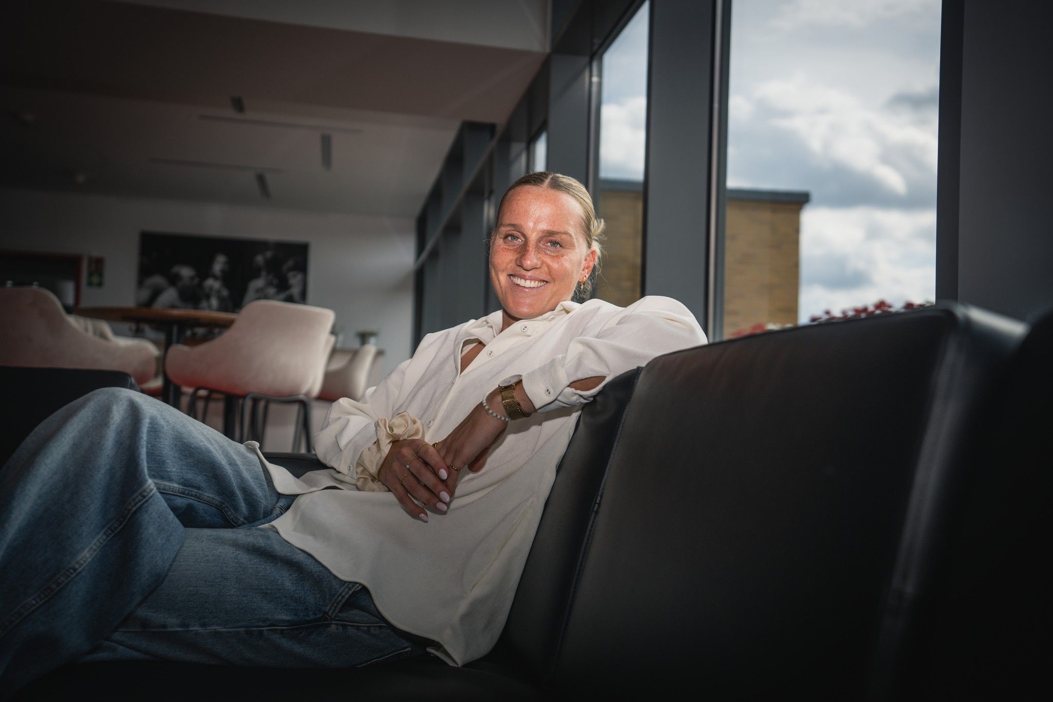Frau mit langen Haare in einem weißen Hemd sitzt entspannt auf einem Sofa in einem modernen Innenraum mit großen Fenstern.