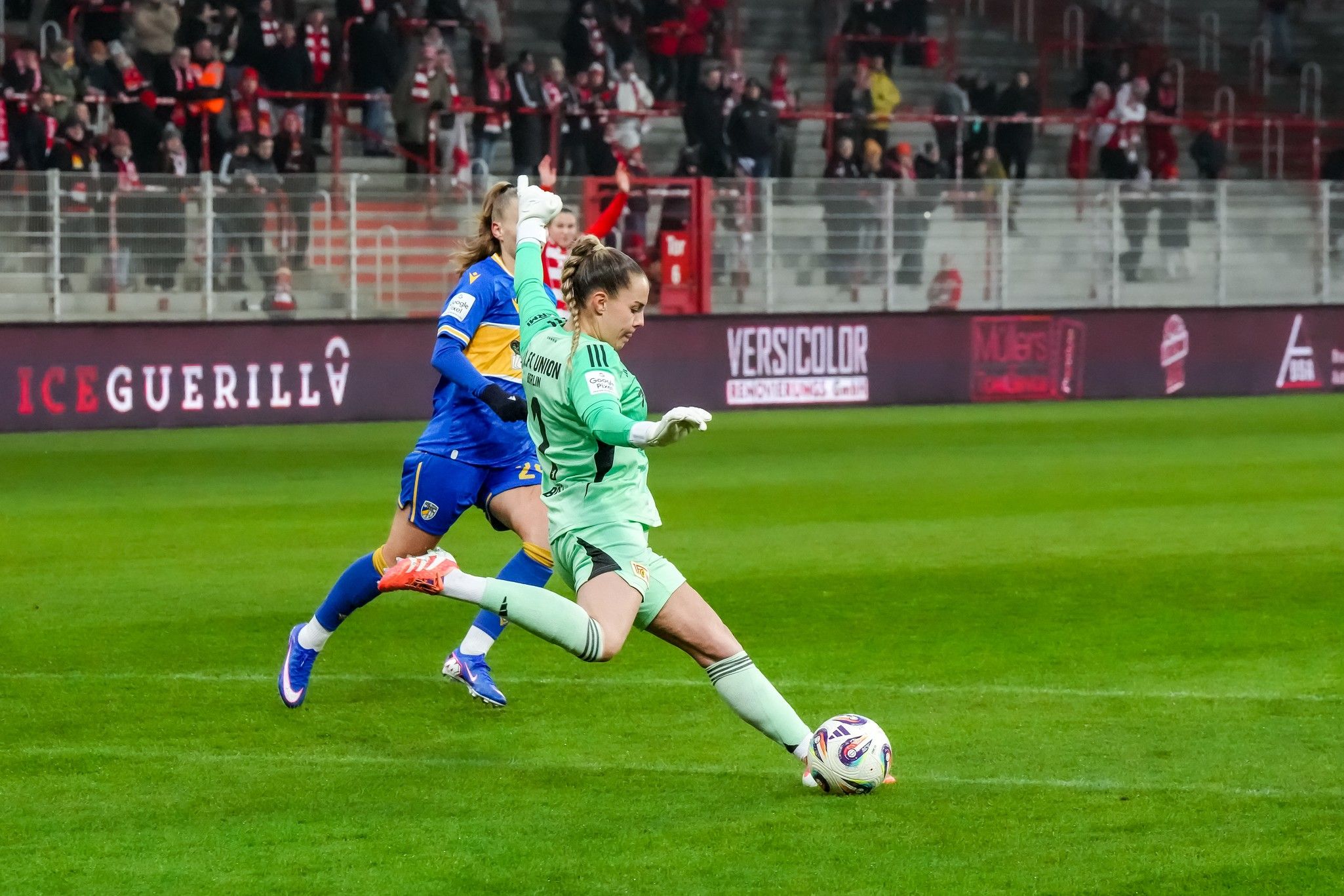 Eine Torhüterin in grüner Ausrüstung wechselt den Ball zu einem Mitspieler, während eine gegnerische Spielerin versucht, ihn zu blockieren.