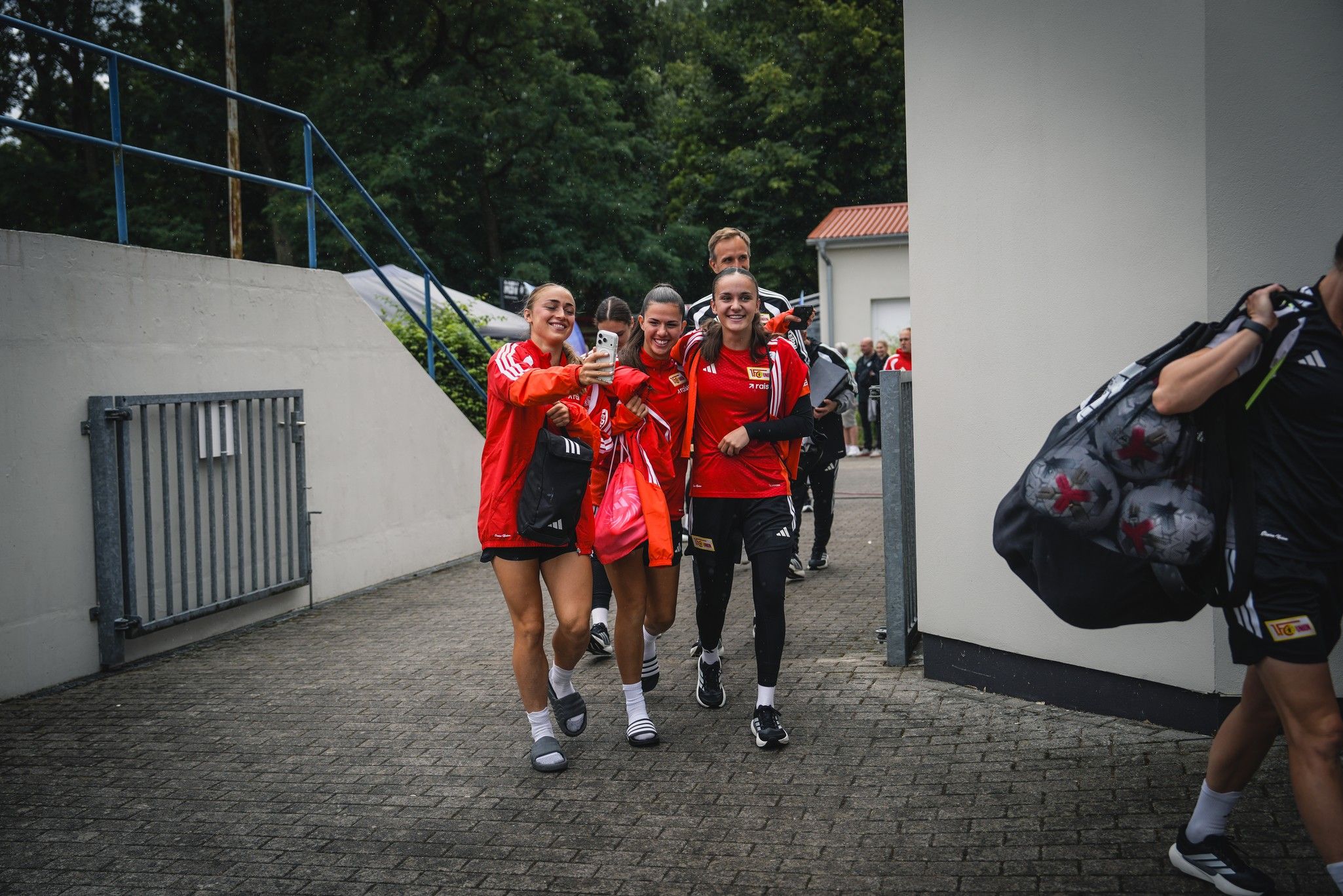 Vier Frauen in roten Sportjacken laufen lächelnd auf einen Eingang zu und halten ein Handy, offenbar beim Aufnehmen eines Selfies.