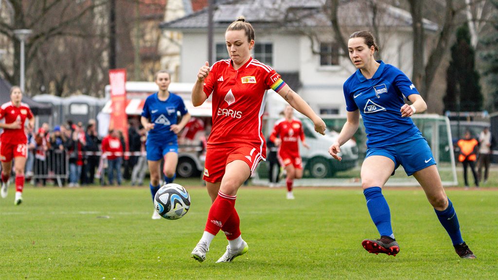 Zwei Frauenfußballspielerinnen in Aktion: eine in rotem Trikot dribbelt den Ball, während eine andere in Blau versucht, den Ball zu erobern.