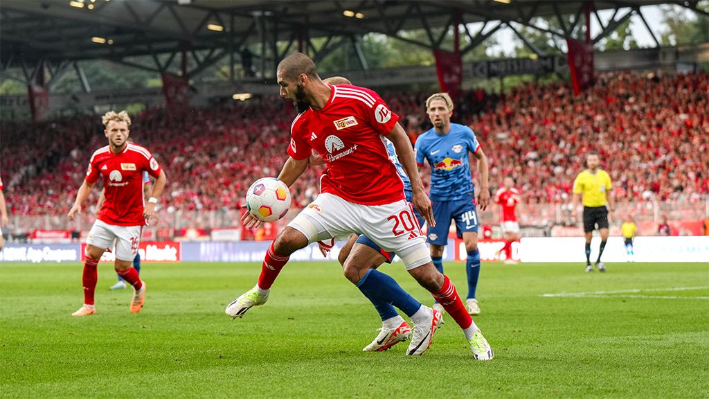 Ein Fußballspieler im roten Trikot dribbelt den Ball, während ein Gegenspieler ihn verfolgt. Im Hintergrund Fans in einem Stadion.