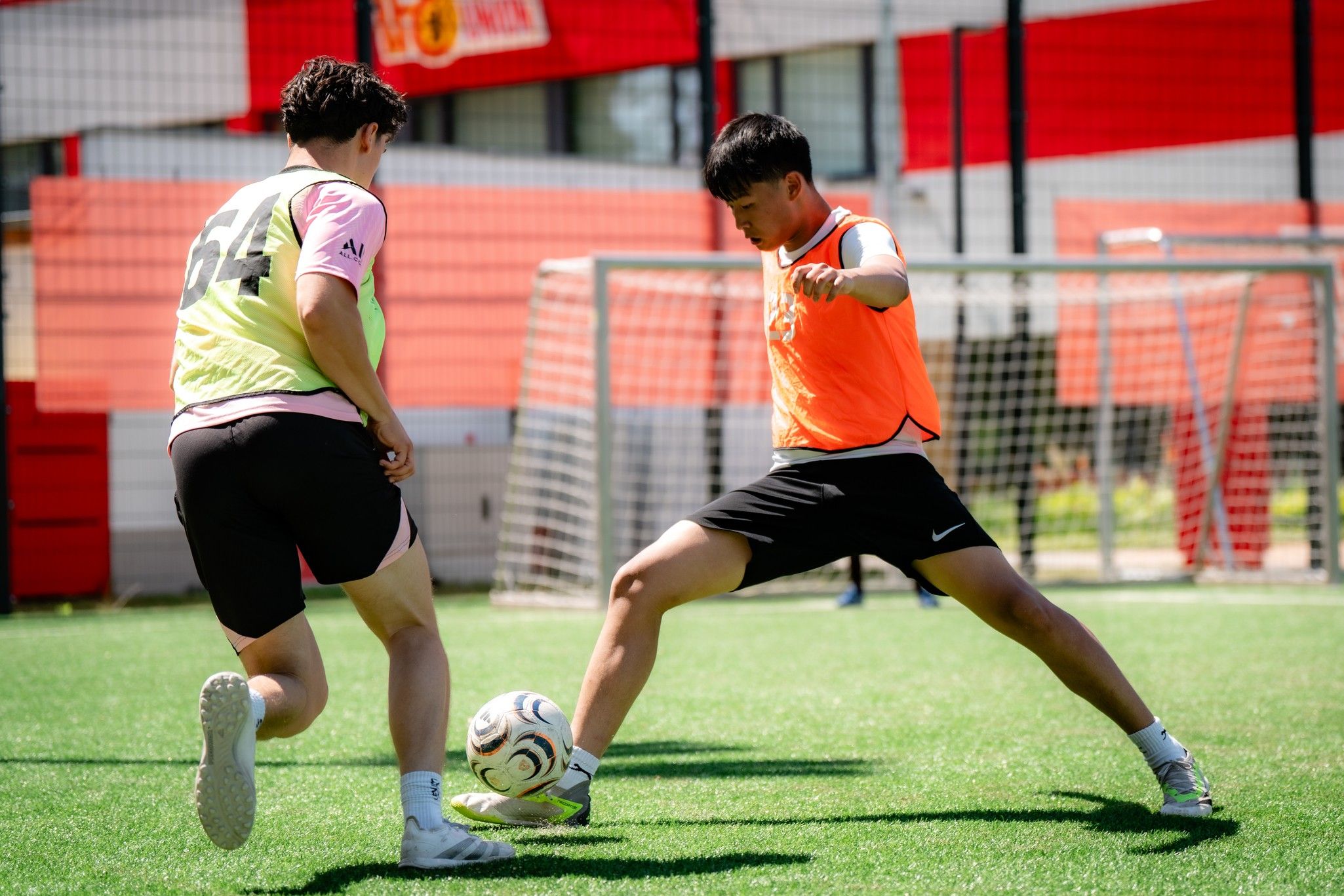 Zwei Fußballspieler in Trainingswesten passen den Ball auf einem Kunstrasenfeld.