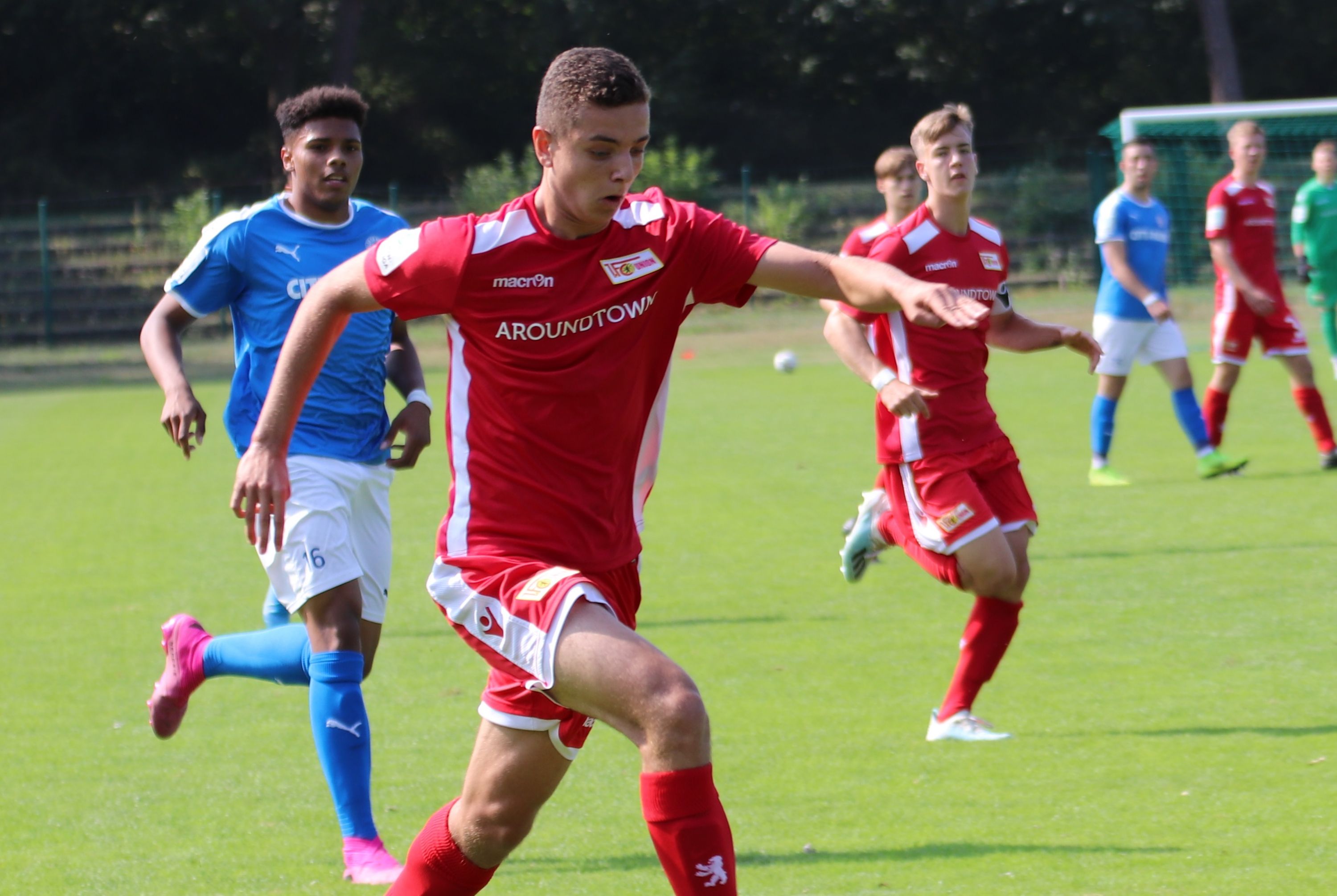 A young soccer player in a red jersey sprints across the field, while other players in blue jerseys stand in the background.