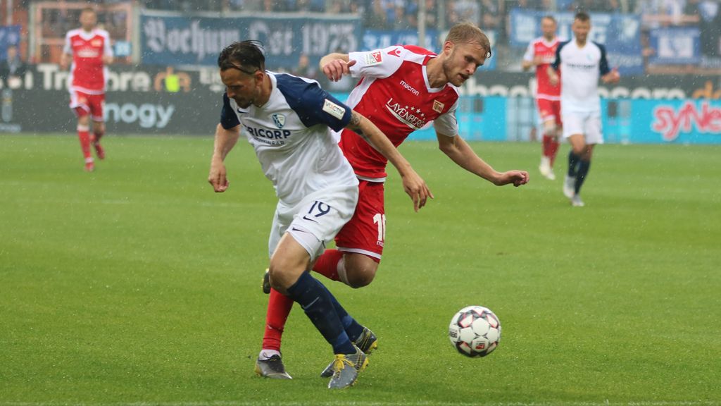 Zwei Fußballspieler kämpfen um den Ball auf einem regennassen Spielfeld. Zuschauer sind im Hintergrund sichtbar.