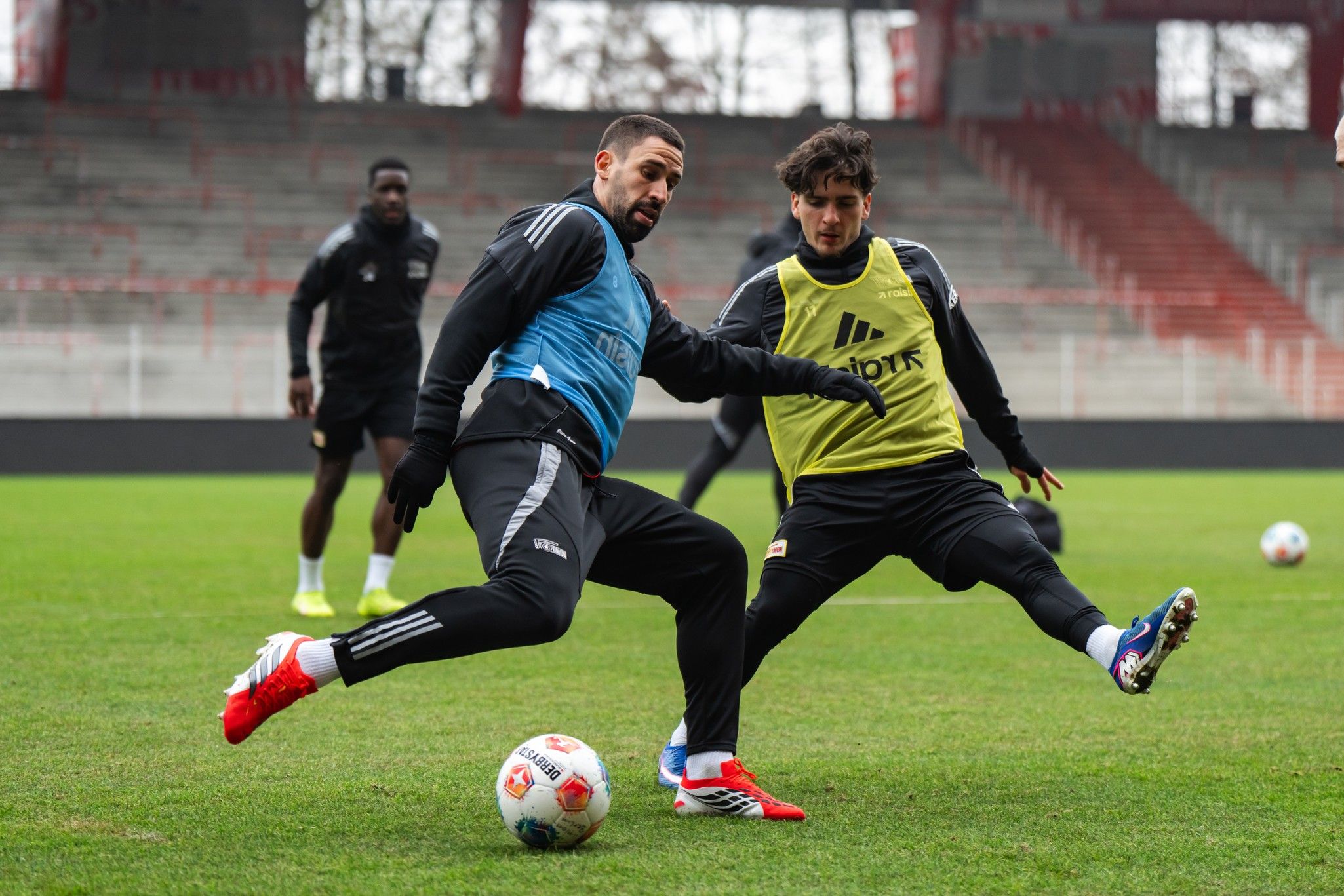 Zwei Fußballspieler im Training, einer mit blauer Weste und der andere mit gelber Weste, kämpfen um den Ball auf einem Rasenplatz.